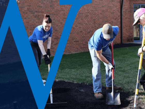 Three Waterford Bank employees in blue shirts working together on a landscaping project, using shovels and rakes to spread mulch near a brick building, with a large translucent blue "W" graphic overlaying part of the image.