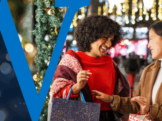 Two women carrying shopping bags and enjoying a holiday outing on a festive city street with lights and decorations in the background.