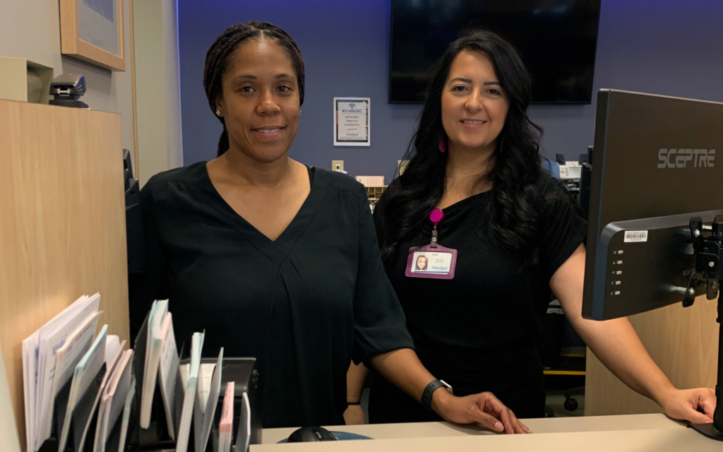 Two bankers stand at a desk with a computer ready to serve a customer.