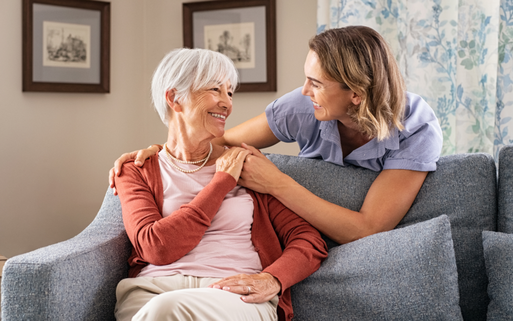 A woman smiling and holding hands with an older woman seated on a couch in a living room.