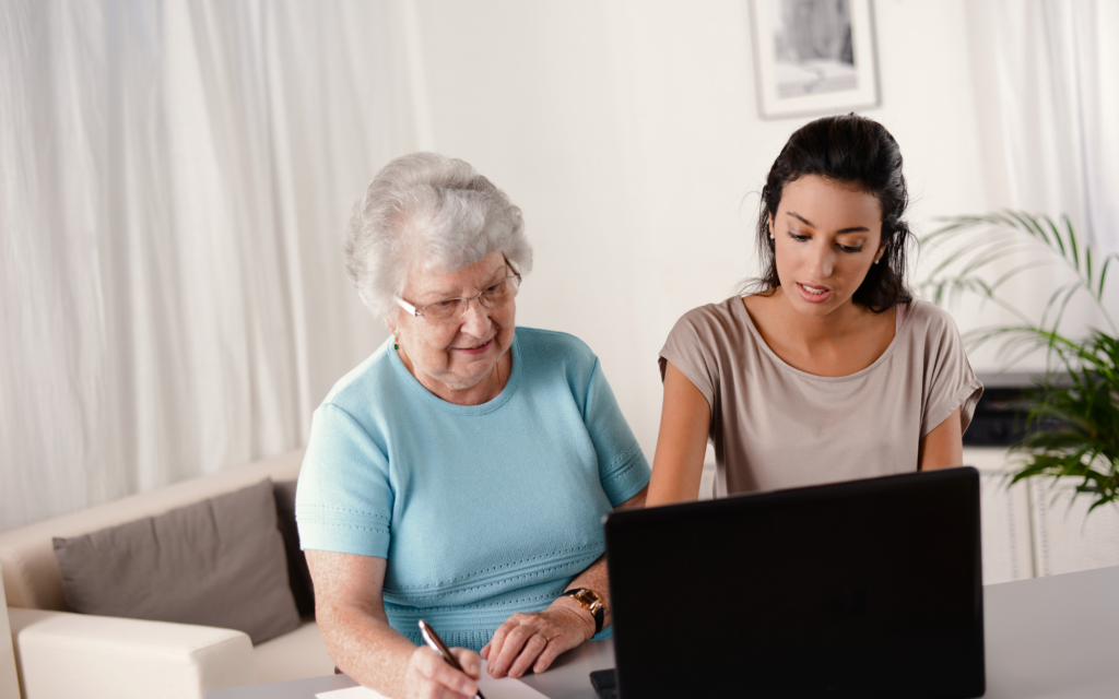 A younger woman helping an older woman review information on a laptop while seated at a table.